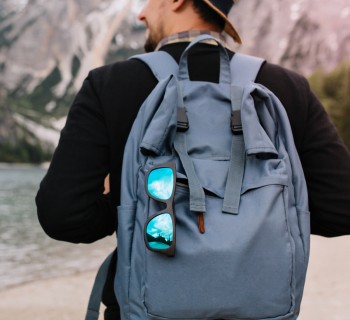 Outdoor portrait from back of male tourist carrying big decorated backpack and walking to mountains in morning. Man wearing black sweatshirt and brown hat travelling around Italy enjoys river view.