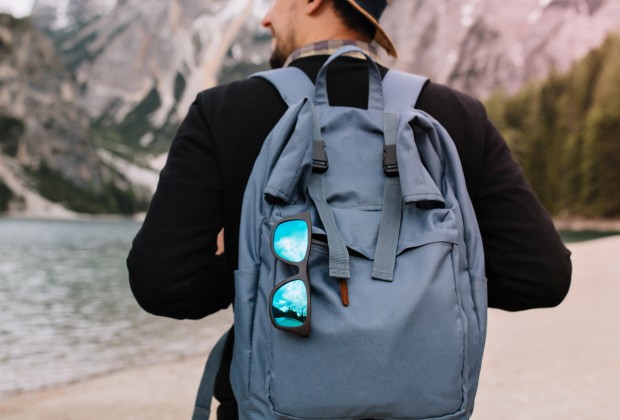 Outdoor portrait from back of male tourist carrying big decorated backpack and walking to mountains in morning. Man wearing black sweatshirt and brown hat travelling around Italy enjoys river view.